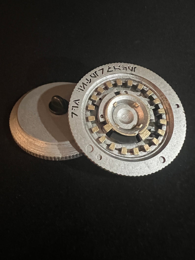 May include: A silver-colored, circular metal object with a complex inner mechanism and gold-colored accents. The object is partially disassembled, revealing the intricate internal design. The rim features etched text. The background is black.