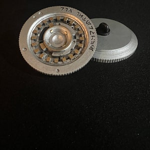 May include: Two-piece, silver-colored metal object with a complex, circular design. The main piece has a central, raised portion surrounded by a series of smaller, rectangular gold-colored elements. The second piece is a flat, silver disc with a black pin.