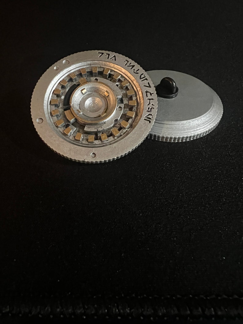 May include: Two-piece, silver-colored metal object with a complex, circular design. The main piece has a central, raised portion surrounded by a series of smaller, rectangular gold-colored elements. The second piece is a flat, silver disc with a black pin.