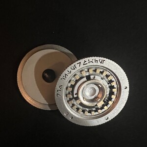 May include: A silver-colored, circular mechanical device with intricate internal gears and a central spindle. The device is partially disassembled, revealing its internal components. The outer ring features etched text. The background is black.