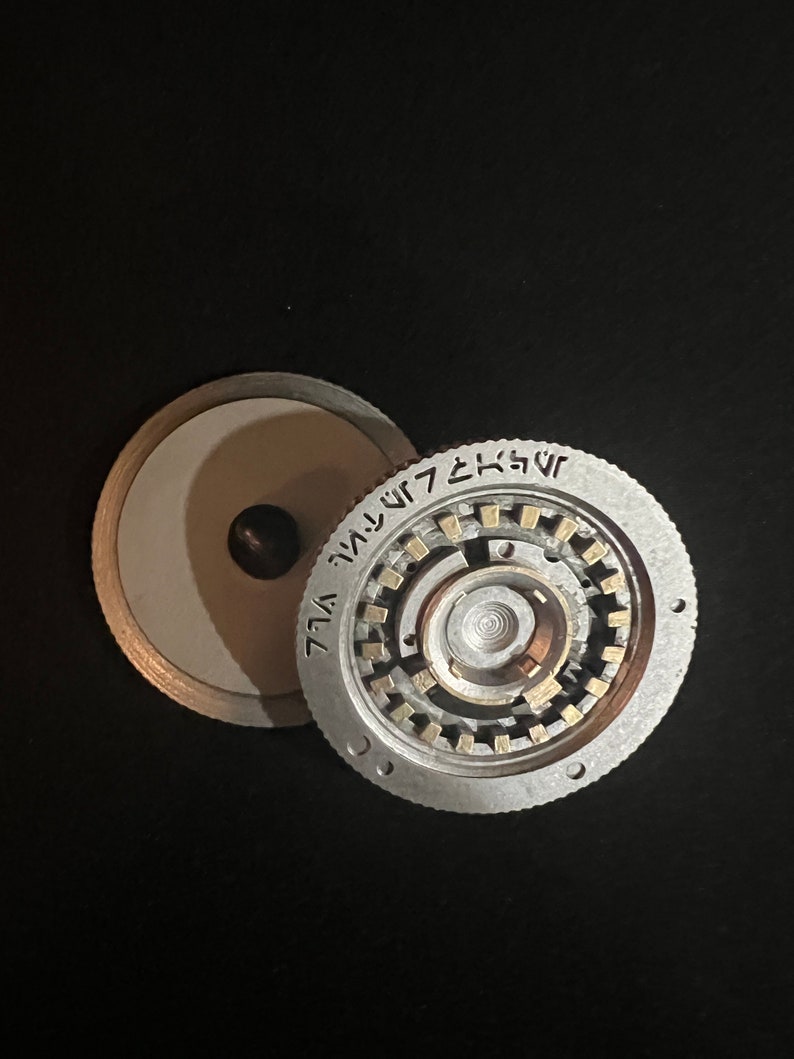 May include: A silver-colored, circular mechanical device with intricate internal gears and a central spindle. The device is partially disassembled, revealing its internal components. The outer ring features etched text. The background is black.