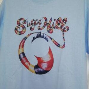May include: Light blue t-shirt with the text "Sugar Hill" in a colorful, patterned font. The design includes a stylized cityscape and a curved, horn-like shape with red, yellow, and purple accents.