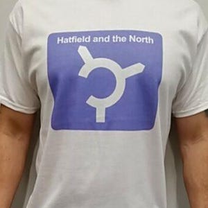 May include: White t-shirt with a blue square graphic featuring a white circular design and the text "Hatfield and the North". The shirt is short-sleeved and appears to be made of cotton.