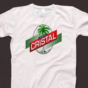 Könnte beinhalten: Weißes T-Shirt mit dem "Cristal"-Logo. Das Logo zeigt eine Palme, rote und grüne Akzente sowie den Text "CERVEZA CRISTAL" und "LA PREFERIDA DE CUBA".
