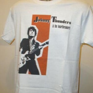 Puede incluir: Camiseta blanca con una imagen en blanco y negro de un guitarrista sobre un fondo naranja. El texto "Johnny Thunders & the Heartbreakers" está impreso en negro encima de la imagen.