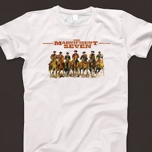 以下が含まれることがあります： 白いTシャツ。7人のカウボーイが馬に乗っているグラフィックと、赤と茶色で「THE MAGNIFICENT SEVEN」の文字がプリントされています。デザインはTシャツの前面中央に配置されています。
