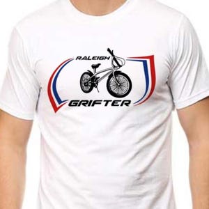 May include: White t-shirt featuring a black and white bicycle graphic with the text "RALEIGH GRIFTER" in a red and blue design. The shirt is made of a soft material and is suitable for casual wear.
