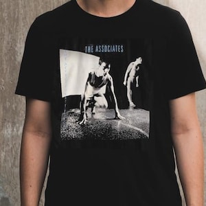 Può includere: T-shirt nero con grafica in bianco e nero di due figure. Il testo "THE ASSOCIATES" è stampato in blu sopra le figure. La maglietta è realizzata in un materiale morbido.