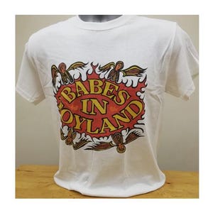 May include: White t-shirt featuring a graphic design with the text "BABES IN OYLAND" in yellow and red, surrounded by flames and angel-like figures. The shirt is displayed on a mannequin.
