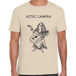 Può includere: T-shirt beige con la scritta "AZTEC CAMERA" sopra un'illustrazione in bianco e nero di una persona che suona la chitarra. L'illustrazione raffigura una persona con i capelli lunghi e un abito fluente.