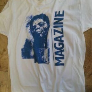 Può includere: T-shirt bianca con grafica blu del volto di una persona e la scritta "MAGAZINE" stampata verticalmente in blu. La maglietta è realizzata in un materiale morbido e confortevole.