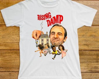 May include: White t-shirt featuring the text "RISING DAMP" in red above a graphic of a man's face and cartoon figures. The graphic includes a hand holding a house and keys. The shirt is made of a soft material.