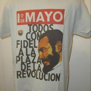 May include: Off-white t-shirt featuring a red rectangular graphic at the top with the text "1° DE MAYO". Underneath is text and a portrait of a man with a beard. The text reads "TODOS CON FIDEL A LA PLAZA DE LA REVOLUCION".
