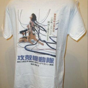 Könnte beinhalten: Weißes T-Shirt mit einer Grafik aus dem Anime Ghost in the Shell. Das Design zeigt eine weibliche Figur, die mit Drähten verbunden ist, mit japanischem Text. Das T-Shirt hat einen Rundhalsausschnitt und kurze Ärmel.