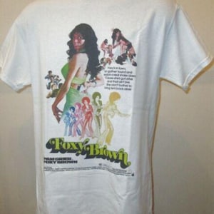 May include: White t-shirt featuring a colorful graphic from the movie "Foxy Brown." The design includes the movie title, a woman in a green dress, and other figures. The shirt has a crew neck and short sleeves.