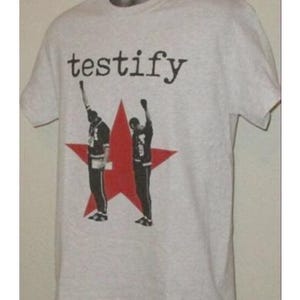 Puede incluir: Camiseta gris claro con la palabra "testify" en negro. Debajo, una estrella roja con dos figuras con los puños en alto. La camiseta es de cuello redondo y manga corta.