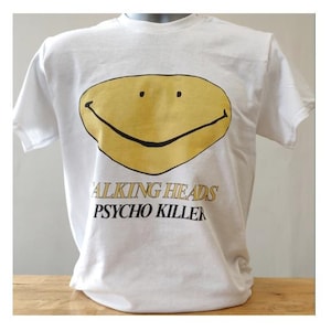 May include: White t-shirt with a large yellow smiley face design. The text "TALKING HEADS PSYCHO KILLER" is printed below the graphic in black and gold. The t-shirt is displayed on a mannequin.