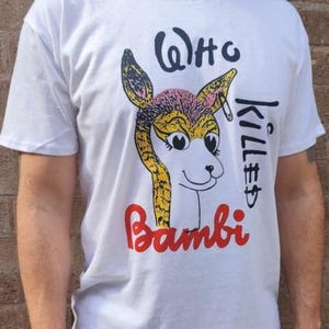 以下が含まれることがあります： 白いTシャツ。漫画の鹿のイラストと「Who Killed Bambi」の文字がプリントされています。鹿は黄色、ピンク、茶色のディテールがあります。Tシャツは半袖のクルーネックです。