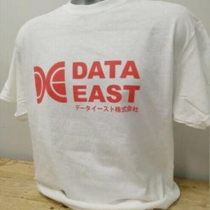Può includere: T-shirt bianca con logo rosso "DATA EAST" e testo giapponese sottostante. La maglietta a maniche corte è realizzata in materiale morbido ed è esposta su un manichino. Un capo semplice e casual.