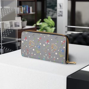 May include: A gray zip-around wallet featuring a repeating pattern of colorful cartoon characters, flowers, and other whimsical elements. The wallet has a gold-colored zipper and a black interior. The design is playful and suitable for everyday use.