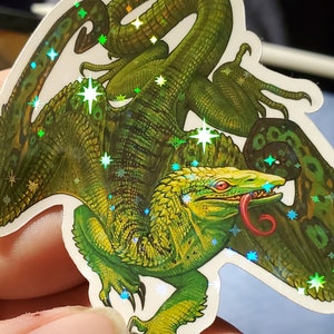 May include: A green and gold iridescent sticker of a mythical creature with a long tail and sharp claws. The creature has a red tongue and is surrounded by stars.