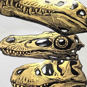 以下が含まれることがあります： 恐竜の頭蓋骨の横顔を描いた、メタリックゴールドとブラックのステッカー3枚。それぞれのステッカーには、先史時代の生物の骨格、歯、眼窩を強調する複雑な線画が施されています。