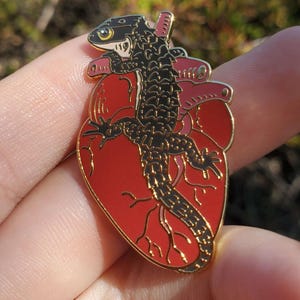 May include: Gold enamel pin featuring a black lizard on a red anatomical heart. The lizard has a yellow eye and is detailed with black scales.