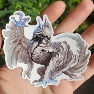 May include: A white and gray detailed sticker of a mythical creature with blue accents. The creature has wings and a long tail. The sticker is on a hand.