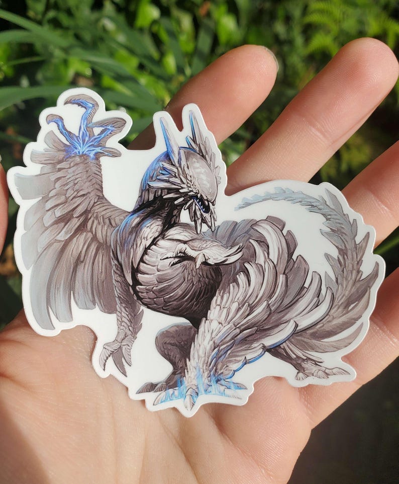 May include: A white and gray detailed sticker of a mythical creature with blue accents. The creature has wings and a long tail. The sticker is on a hand.