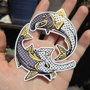 May include: A die-cut sticker featuring two stylized fish arranged in a circular formation. The fish are illustrated with a black and white polka dot pattern, accented with solid yellow fins and tails, and feature bold black outlines.