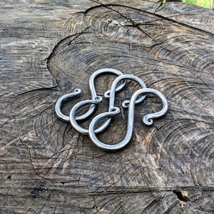 May include: Three silver metal hooks with a decorative swirl design. The hooks are arranged in a pile on a wooden surface.