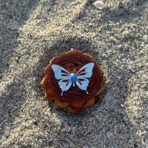 May include: A circular brown wooden slice featuring a silver-toned butterfly cutout ornament centered on its surface. The butterfly has an iridescent blue oval cabochon stone set in its middle, creating a contrast against the textured wood grain.