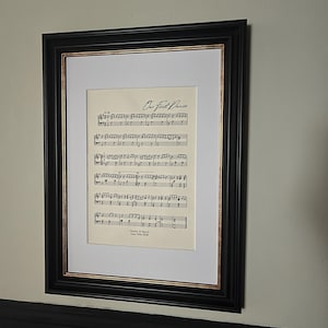 May include: A framed print of sheet music, titled "Our First Dance". The black frame has a white mount with a gold inner edge, accentuating the musical notes. A decorative piece of wall art, perfect for a music lover's home.