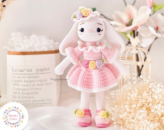 Puede incluir: Una muñeca de conejo de crochet que lleva un vestido rosa con un borde de encaje blanco y una corona de flores. La muñeca tiene una cara blanca con ojos negros y una nariz rosa. La muñeca está de pie sobre una superficie blanca con una bolsa de papel blanca y marrón en el fondo.