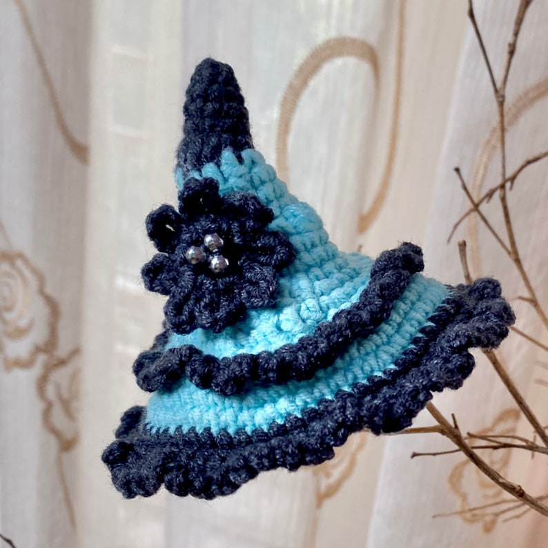 May include: A blue and grey crocheted witch hat with a black flower and silver beads.