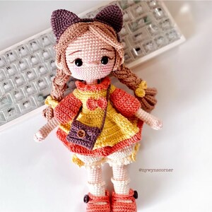 May include: A crocheted doll with brown hair, wearing a yellow and orange dress with a brown purse. The doll is wearing brown shoes and has a pink bow in her hair.