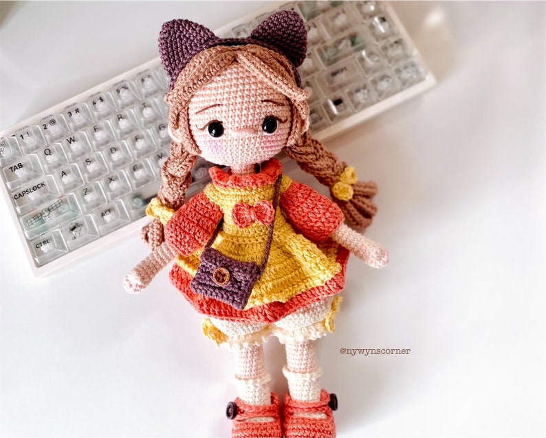 May include: A crocheted doll with brown hair, wearing a yellow and orange dress with a brown purse. The doll is wearing brown shoes and has a pink bow in her hair.