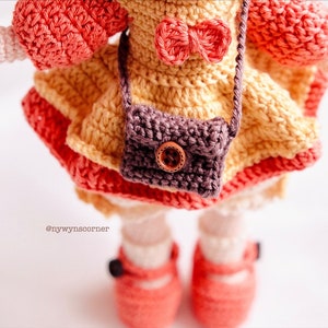 May include: A crocheted doll wearing a yellow dress with a brown purse and a red bow. The doll is wearing red shoes and has pink arms.