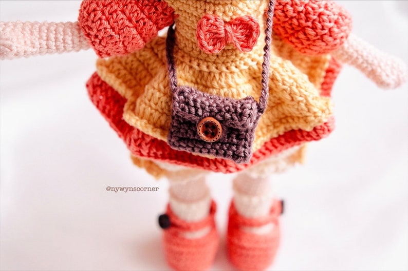 May include: A crocheted doll wearing a yellow dress with a brown purse and a red bow. The doll is wearing red shoes and has pink arms.