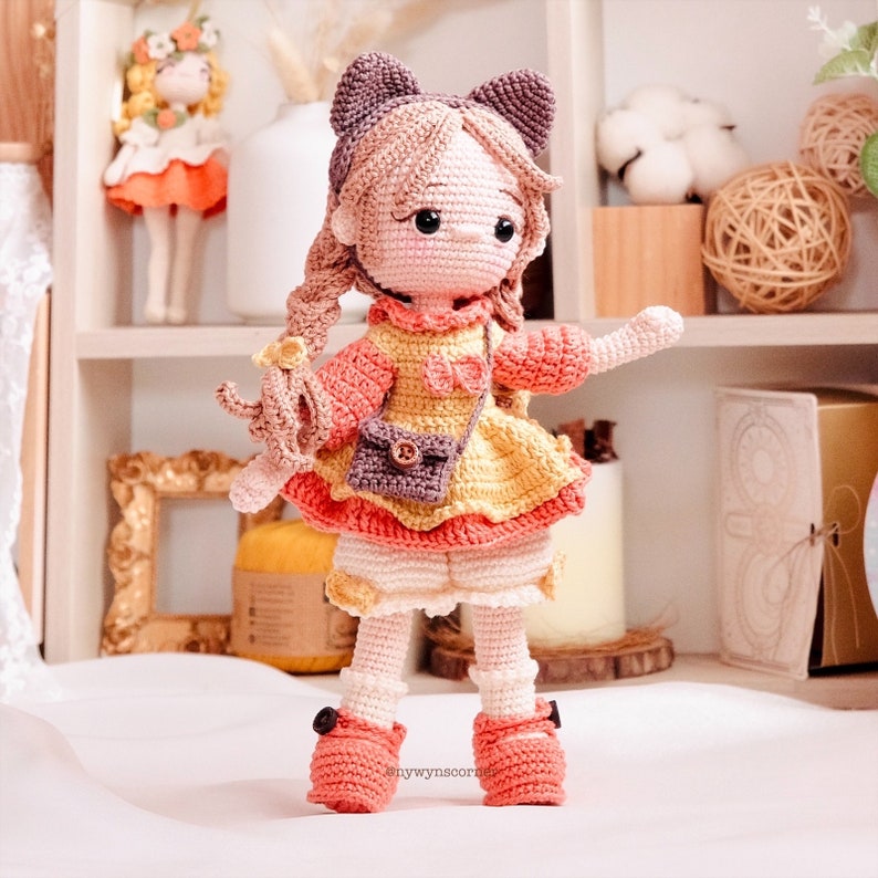 May include: A crocheted doll with brown hair, wearing a yellow and pink dress, a brown purse, and pink shoes. The doll is standing on a white surface.