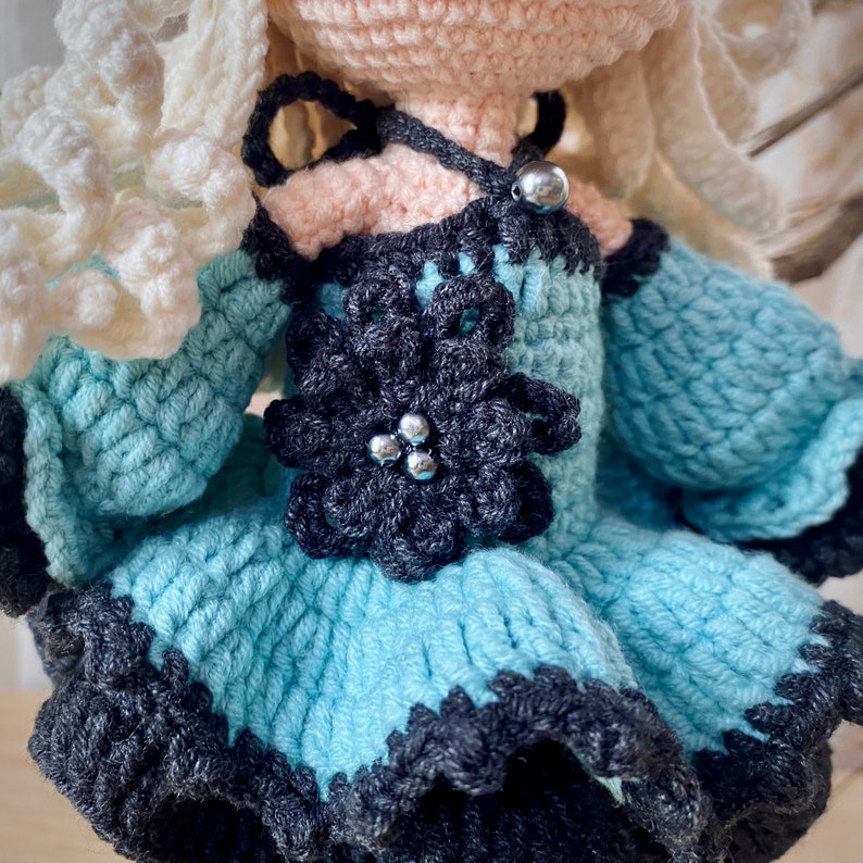 May include: A crocheted doll wearing a light blue dress with a black flower and silver beads. The doll has white yarn hair and a pink face.