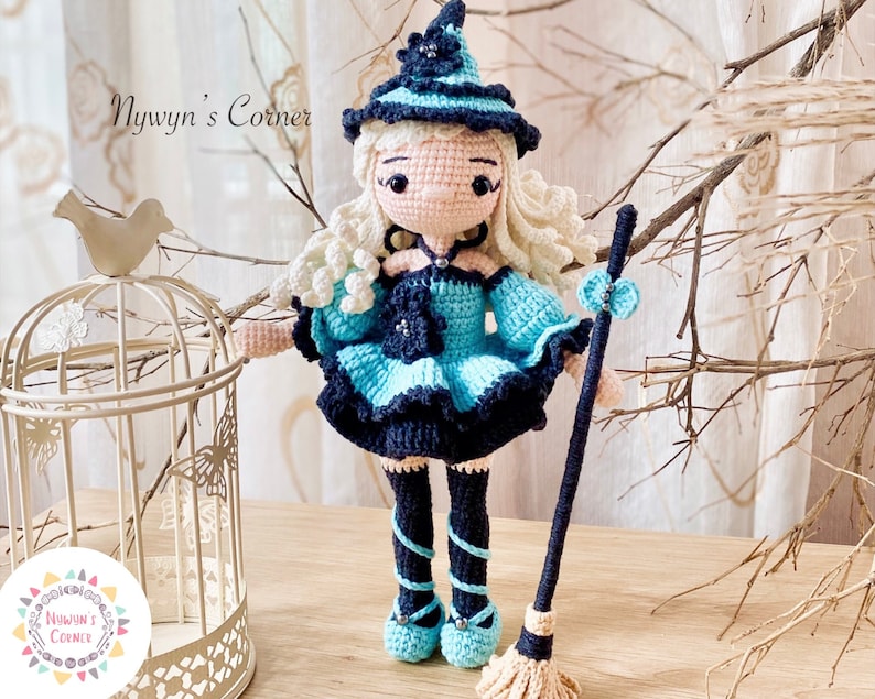 May include: A crocheted witch doll with blonde hair, wearing a blue and black dress and a witch hat. She is holding a broom in her right hand. The doll is standing on a wooden surface.