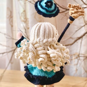 May include: A crocheted doll with long blonde hair wearing a blue and black dress and holding a brown broom. The doll is wearing a blue and black witch hat.