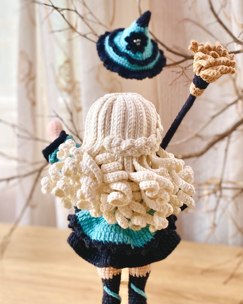 May include: A crocheted doll with long blonde hair wearing a blue and black dress and holding a brown broom. The doll is wearing a blue and black witch hat.