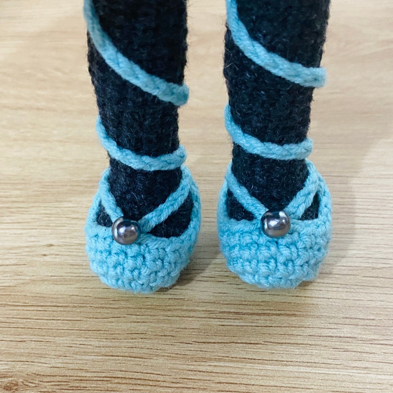 May include: A pair of light blue crocheted ballet flats with black beads on the front. The flats are worn by a doll with black crocheted legs.