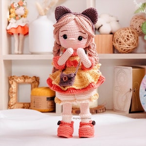 May include: A crocheted doll with brown hair, wearing a yellow and pink dress, a brown purse, and pink shoes. The doll has cat ears and is holding its hands together.