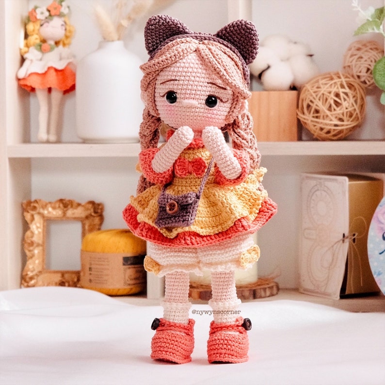 May include: A crocheted doll with brown hair, wearing a yellow and pink dress, a brown purse, and pink shoes. The doll has cat ears and is holding its hands together.