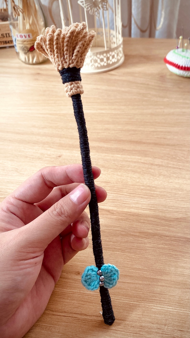 May include: A small, crocheted broom with a brown handle and a blue bow. The broom is made of beige yarn and has a black band around the handle.