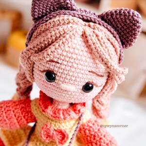May include: A crocheted doll with pink hair, brown eyes, and a pink and yellow dress. The doll is wearing a purple headband with cat ears.