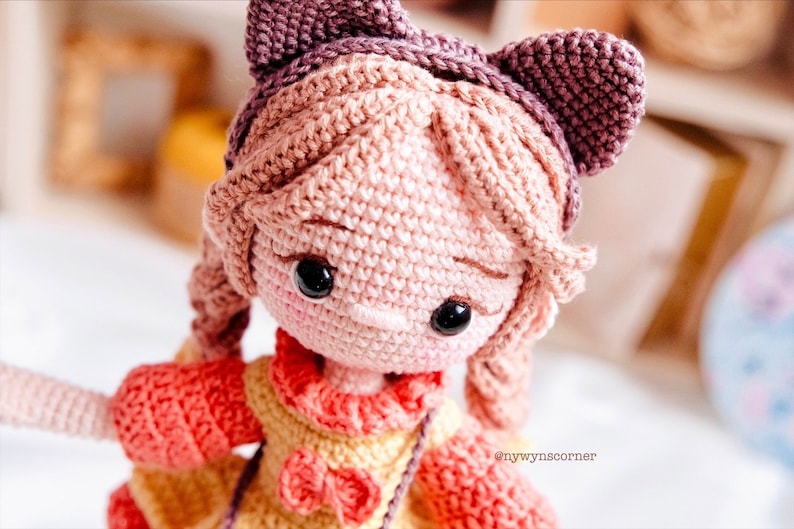 May include: A crocheted doll with pink hair, brown eyes, and a pink and yellow dress. The doll is wearing a purple headband with cat ears.